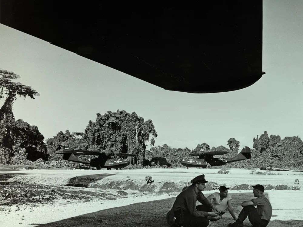 Three men sit under the shade of a massive airplane wing; one of the men is wearing an officer's hat. Two huge aircraft are parked in the background, both are painted black. Behind them is a lush island landscape. The photo has been given a green tint.