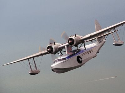 Silver twin engine flying boat over a vast ocean. A small boat leaves it's wake below.